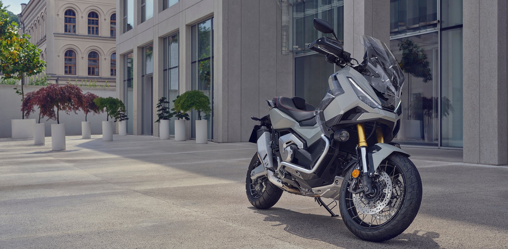 Honda X-ADV argent stationné sur une place urbaine devant une architecture de verre